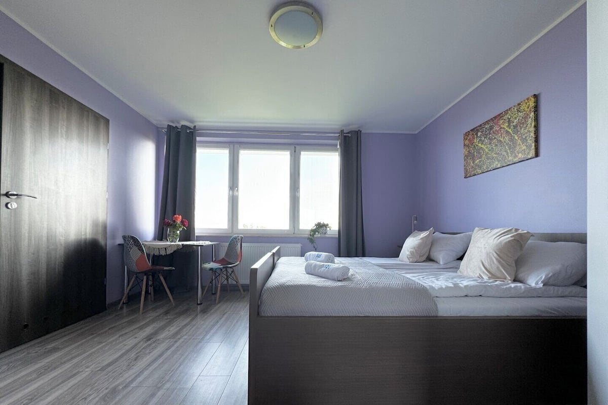 A modern hotel room with a large bed, a small desk with a chair, a window with curtains, and a ceiling light.