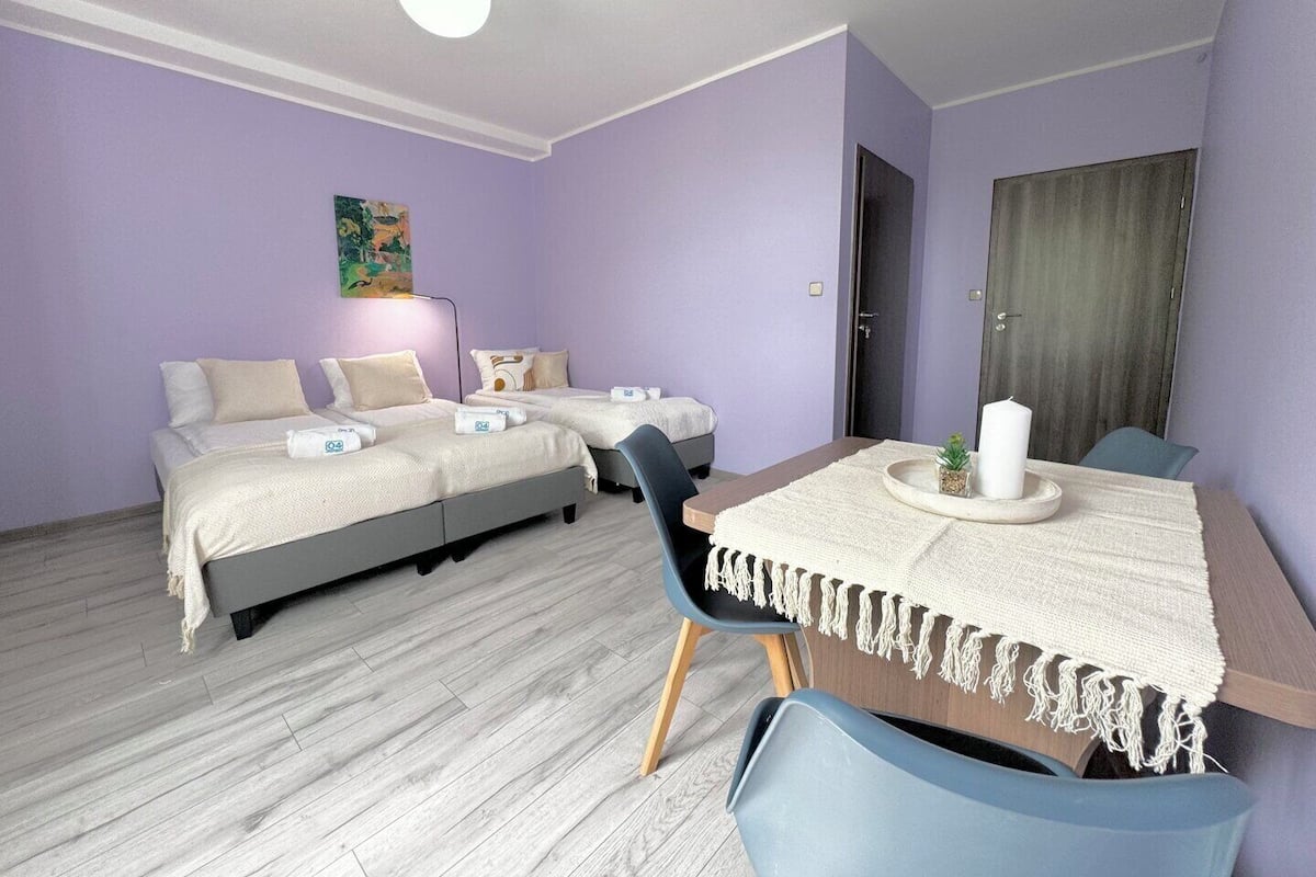 A room with two beds, a dining table with chairs, a small plant, and a painting on the wall.
