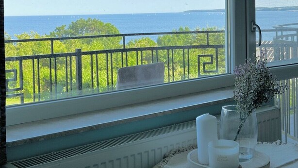 A room with a view of the sea through a window with a black metal railing. There is a radiator below the window and a small table with candles and a vase of flowers on it. - Pensjonat Błękitna Przystań (Puck)