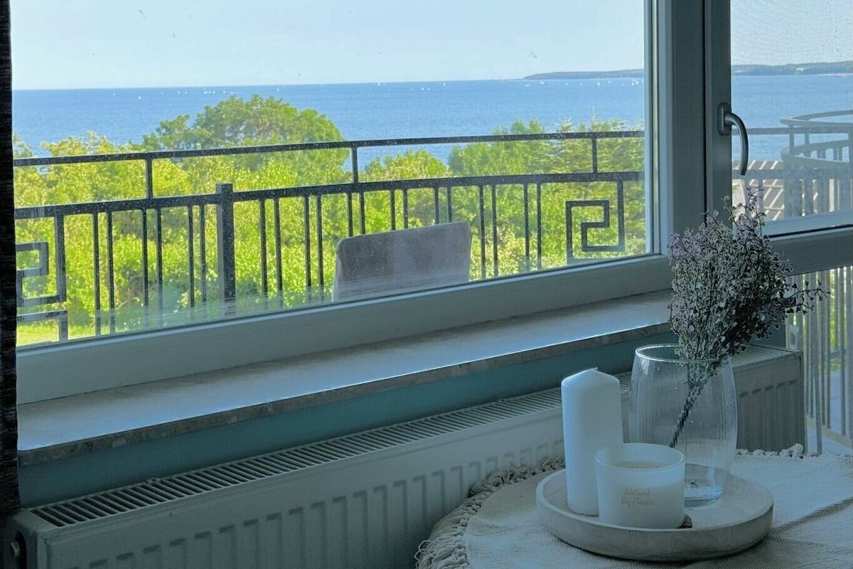 A room with a view of the sea through a window with a black metal railing. There is a radiator below the window and a small table with candles and a vase of flowers on it.