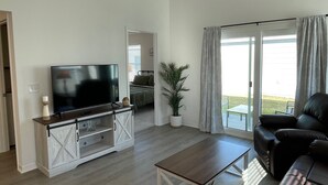 A modern living room with a flat-screen TV on a wooden stand, a black leather sofa, a wooden coffee table, and a large window with curtains. - Cozy Retreat Near Sawgrass Market & Brownwood Paddock Square! (Wildwood)