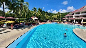 Una gran piscina rodeada de sillones de descanso y sombrillas, con palmeras y un edificio al fondo.