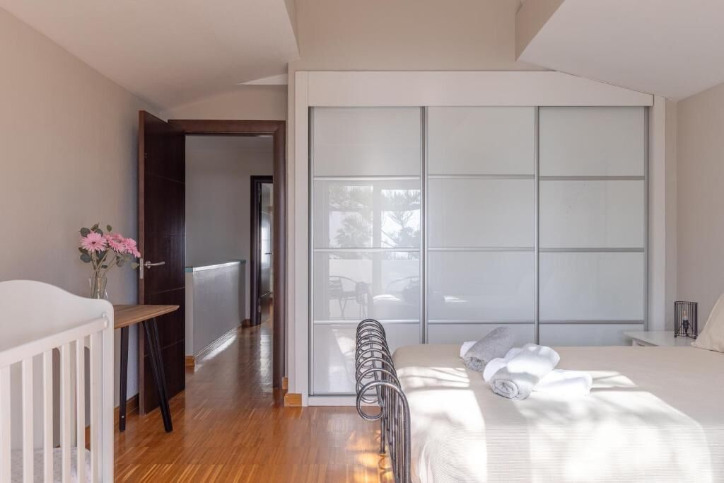 A modern bedroom with a large bed, a wooden floor, a white crib, and a sliding glass door leading to a balcony.