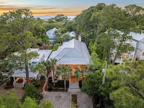A coastal house with a prominent gabled roof, surrounded by dense greenery and other residential buildings.