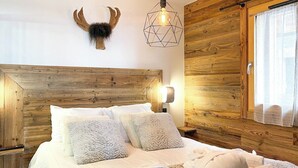 A neatly made bed with white linens and fluffy pillows, a wooden headboard, a wall-mounted deer head, and a geometric pendant light. - Apartment Lumikki |  Private hot tub and sauna (Les Gets)