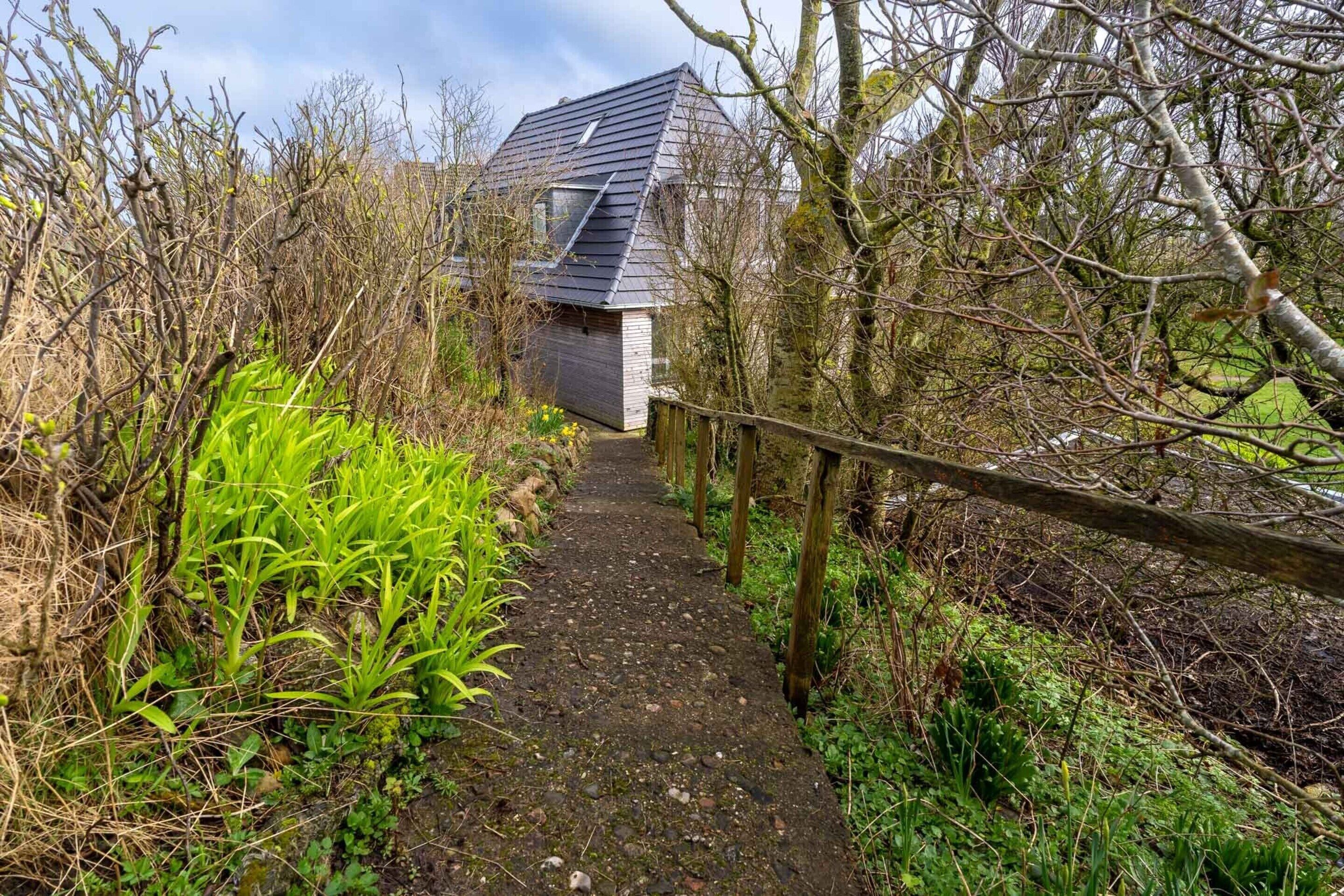 Un sentier étroit et non pavé mène à une maison dont le toit est en pente, entourée d’une végétation dense et d’une clôture en bois.
