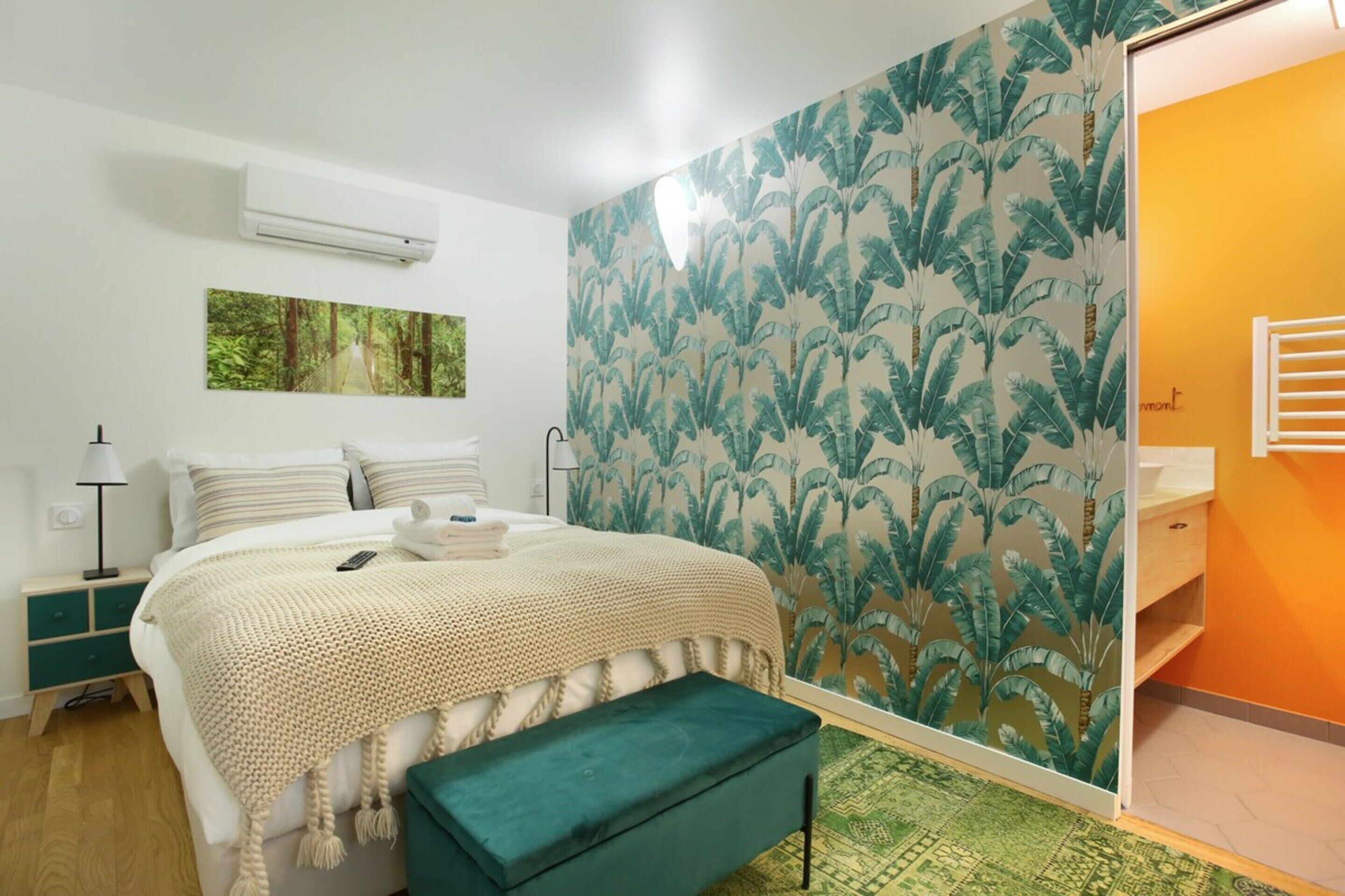 Un dormitorio con cama, una banca, mesita de noche, lámpara y un baño con toallero.