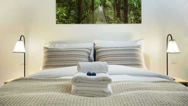 Un letto rifatto con cura, un asciugamano piegato e due bicchieri, affiancati da due lampade e un arazzo a tema foresta.