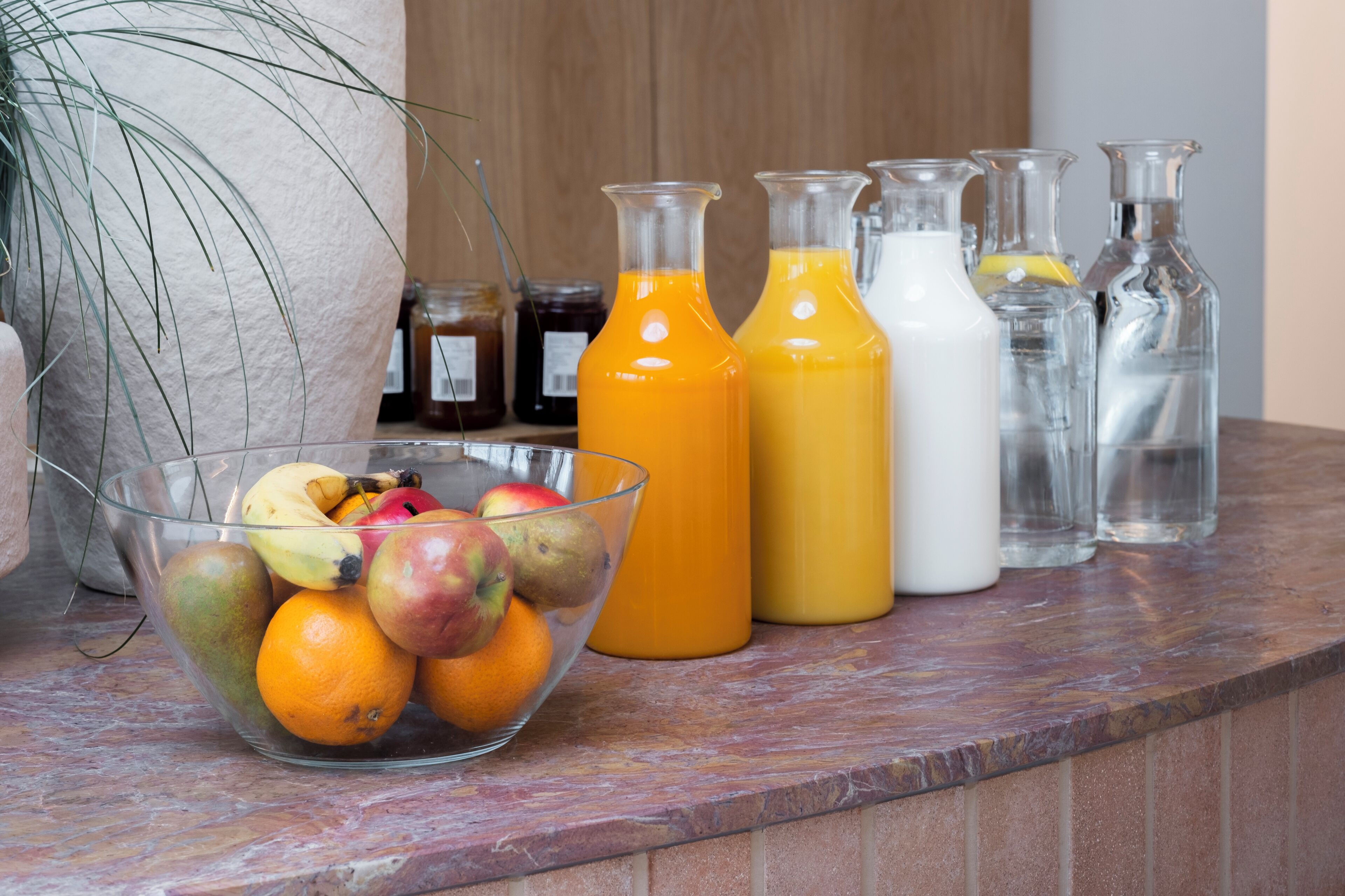 A kitchen counter with a glass bowl of fruit, several glass bottles of orange juice, and a potted plant.