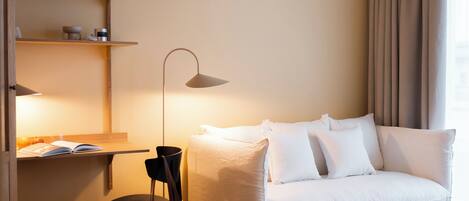 A modern hotel room with a white sofa, a small side table, a lamp, a chair, a woven basket, and a wooden floor.