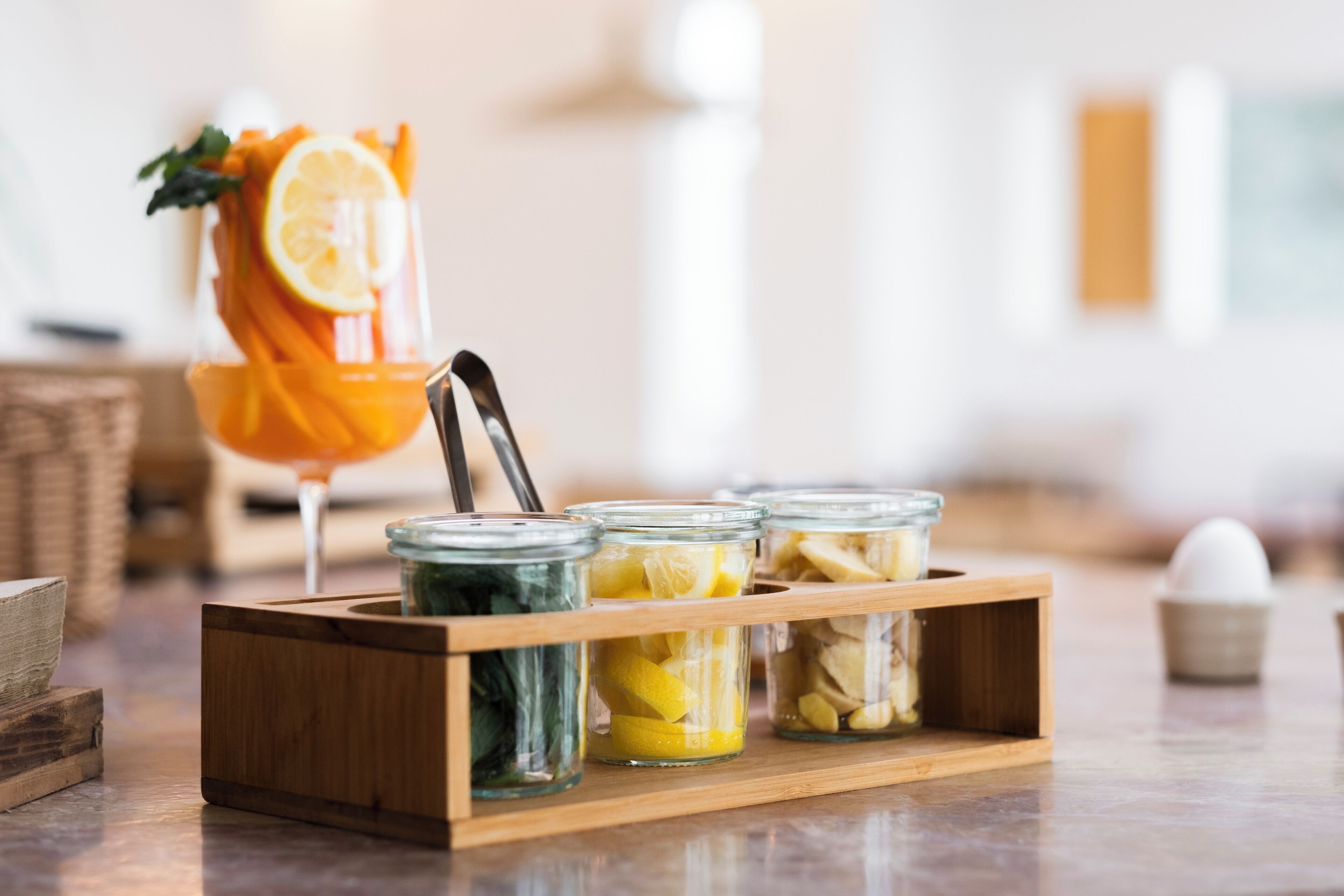 A wooden tray with glass jars containing preserved lemons and oranges, and a glass with a lemon slice garnish.