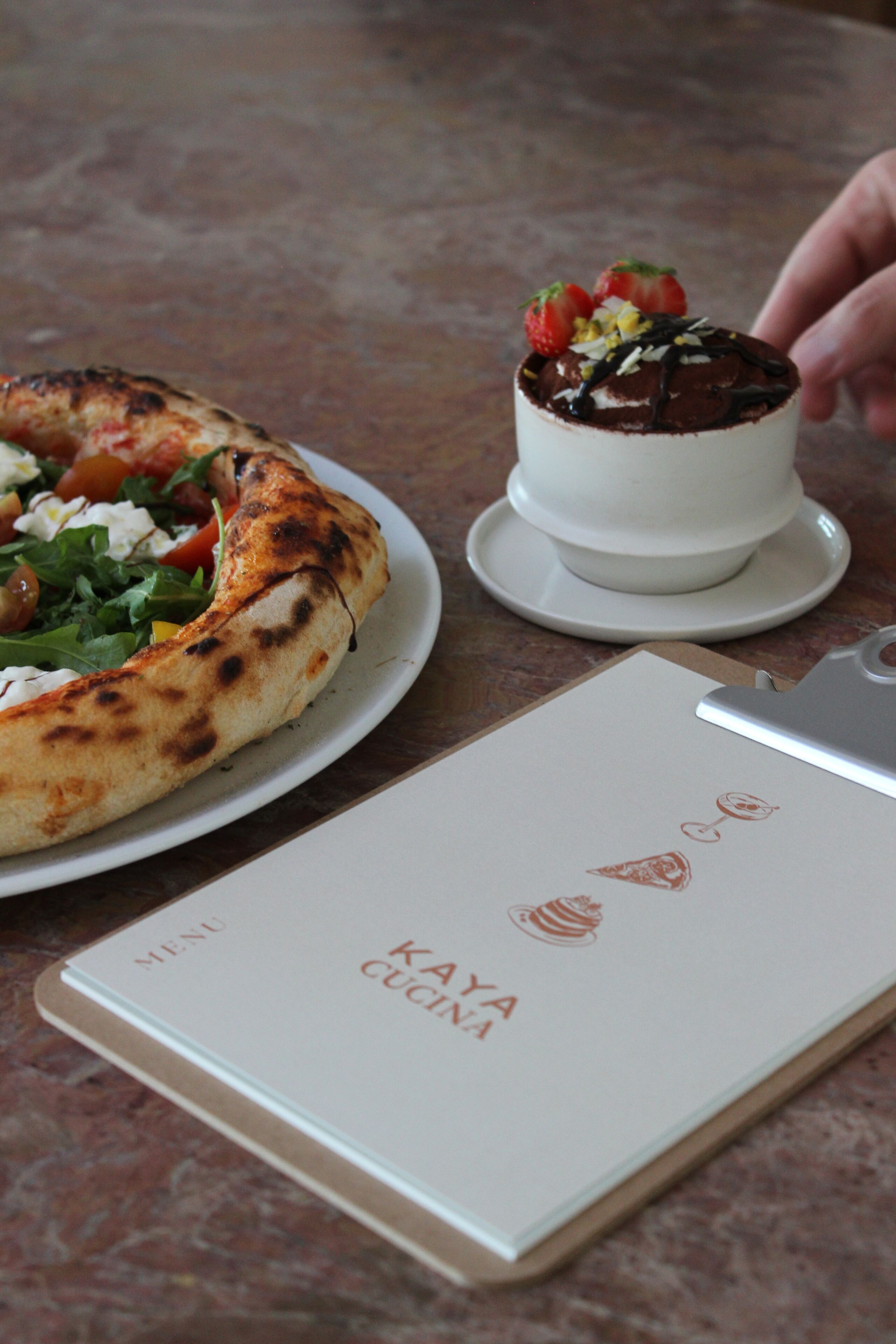 Eine Speisekarte mit der Aufschrift "KAYA CUCINA" auf einem Tisch mit Pizza und Dessert.