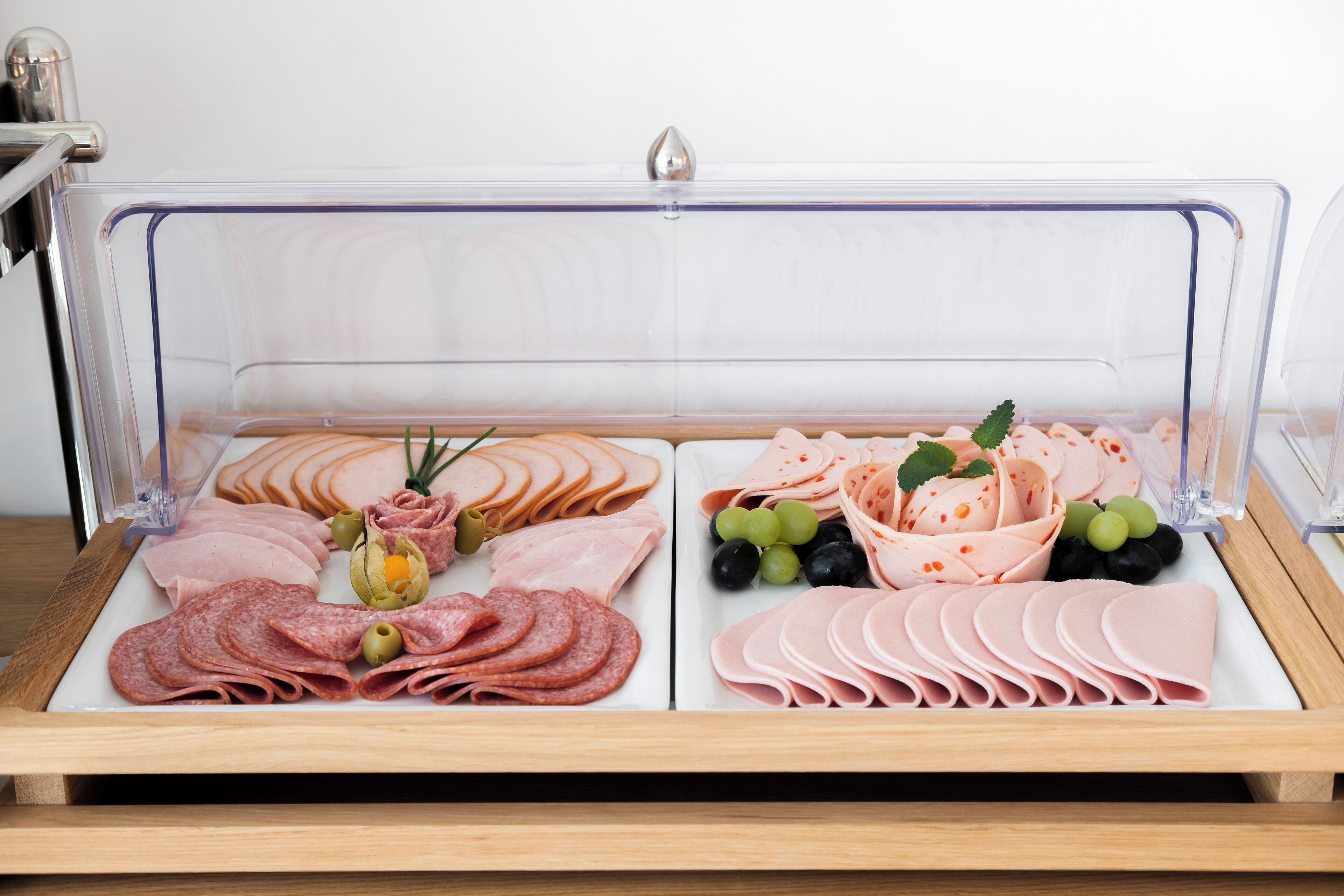 A clear glass display case with various types of sliced meats and cheeses, including salami, ham, and cheese slices, arranged neatly.
