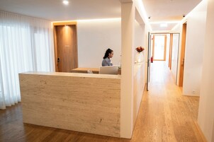 Zona de recepción con un mostrador de madera, una persona sentada frente a un ordenador portátil y un pasillo que conduce a una salida.