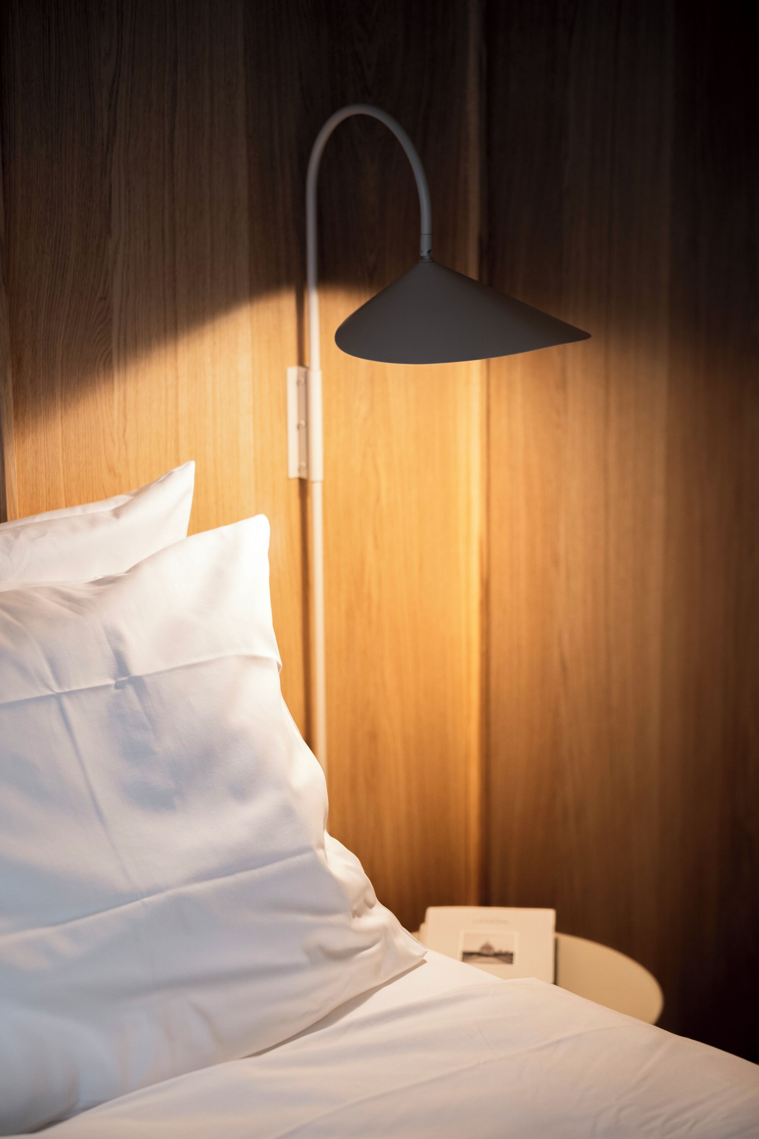 A neatly made bed with white linens and pillows, a modern wall-mounted lamp, and a small bedside table with a book.