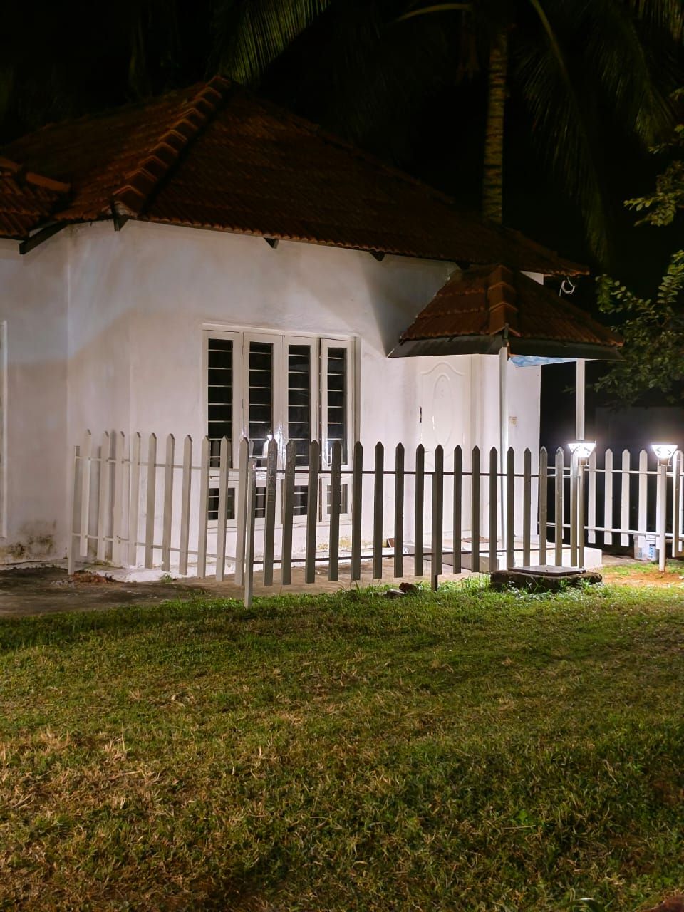 A small, single-story building with a white exterior and a red-tiled roof. It features a white picket fence and a porch with a white railing.