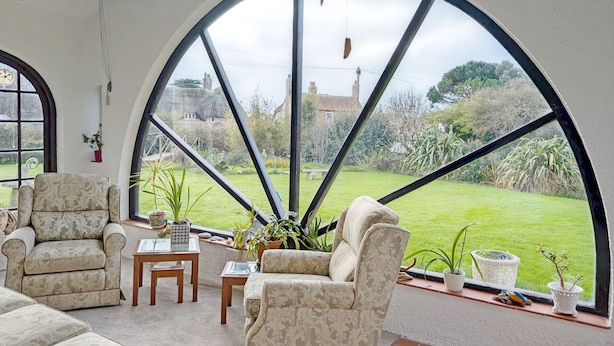 A spacious room with a large arched window, a comfortable seating area with a sofa and armchairs, a small table, and various potted plants.