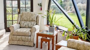 A cozy room with a patterned armchair, a small wooden table, and a view of a garden through a large arched window.