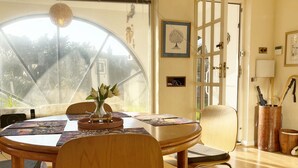 A dining area with a round table, chairs, a vase with flowers, and a view through a large window.