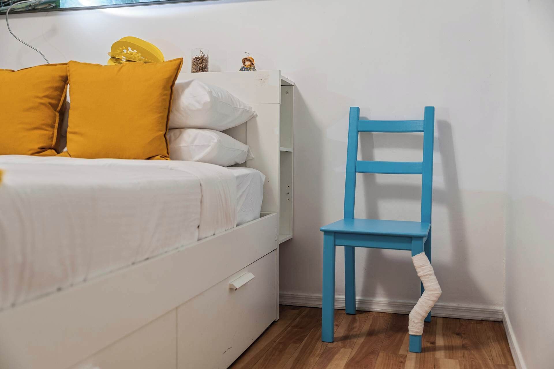 A bedroom with a bed, a white dresser, a blue chair, and a small shelf with decorative items.