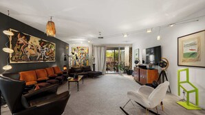Een moderne woonkamer met een bank, een witte fauteuil en een televisie. Op de muren hangen schilderijen en er is een groot raam.