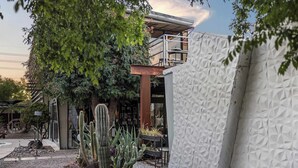 Een modern gebouw met een geometrisch gevelontwerp, een terras op het dak en diverse cactussen en planten in de omliggende tuin.