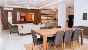 A modern living room with a wooden dining table, grey upholstered chairs, a white sofa, and a television displaying a music album.