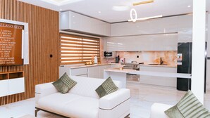 A modern kitchen with white cabinets, a built-in oven, and a refrigerator. There is a white sofa with green cushions in the living area.