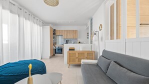 A modern living room with a grey sofa, a blue accent chair, and a white table with a decorative item. A kitchen with wooden cabinets and a dining area is visible in the background. - Le Nid Bleu - Entre Mer et Montagne (Grosseto-Prugna)
