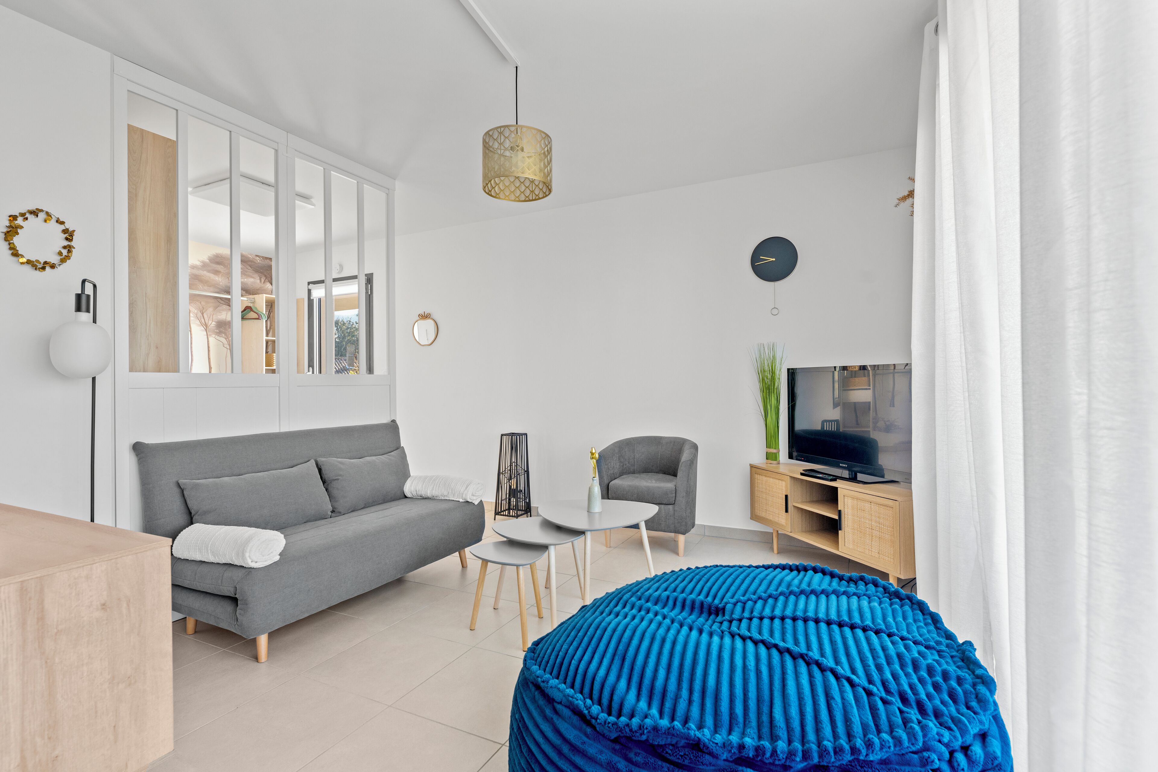 A modern living room with a grey sofa, a blue pouf, a small white table, and a TV on a wooden cabinet.