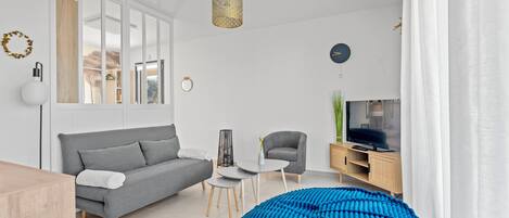 A modern living room with a grey sofa, a blue pouf, a small white table, and a TV on a wooden cabinet.