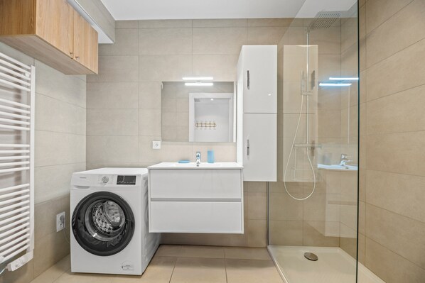 Une petite buanderie comprenant un lave-linge, un évier et une douche fermée par une paroi en verre.