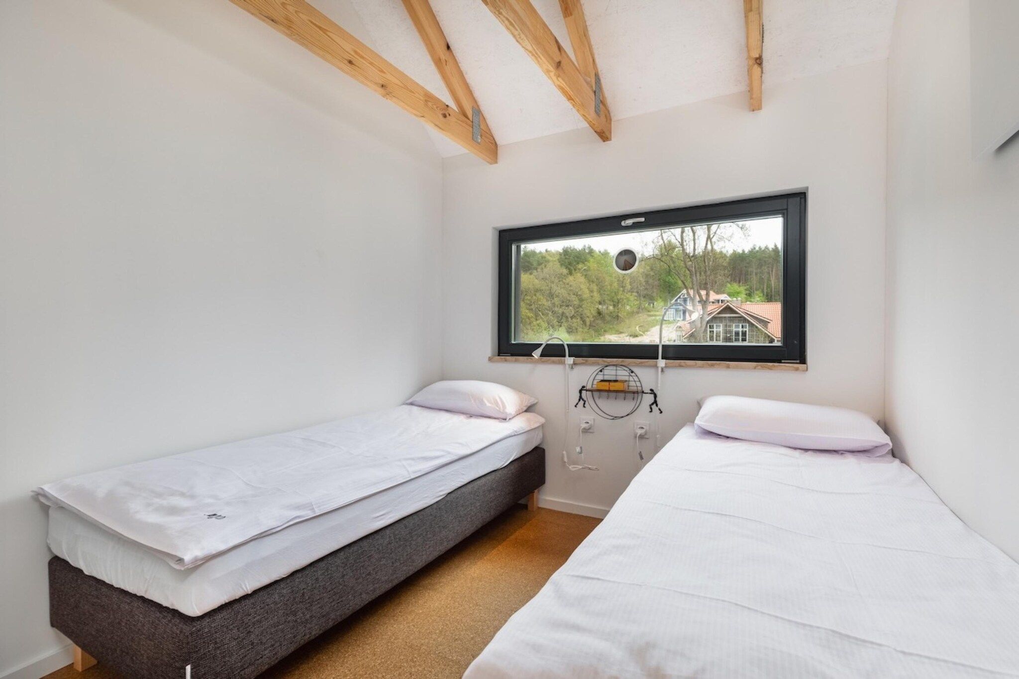 A small room with two single beds, a window with a view of houses, and exposed wooden beams on the ceiling.