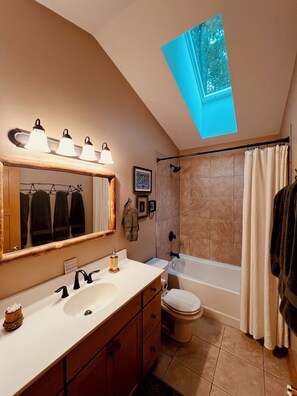 A modern bathroom with a skylight, a bathtub, a toilet, and a vanity with a mirror and lights.