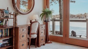 Una habitación con un aparador de madera, una silla, una lámpara, un espejo y un ventanal grande con vista al agua.