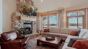 Un salón acogedor con chimenea de piedra, un sofá de cuero, una mesa de centro de madera y ventanas amplias con vistas al exterior.