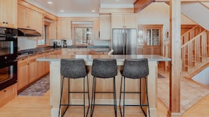 Una cocina moderna con armarios de madera, una isla central con taburetes de bar, electrodomésticos de acero inoxidable y una escalera de madera.
