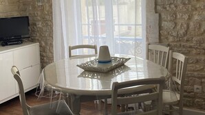 Un comedor con una mesa redonda, sillas blancas, un televisor sobre un mueble y una pared de piedra.
