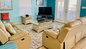 A living room with beige leather sofas, a glass coffee table, a flat-screen TV, and a ceiling fan.