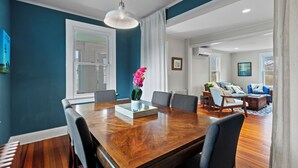Ein Essbereich mit Holztisch, Stühlen und einer Vase mit Blumen. Im Hintergrund befindet sich ein Fenster mit Vorhängen, ein Sofa und ein Couchtisch.