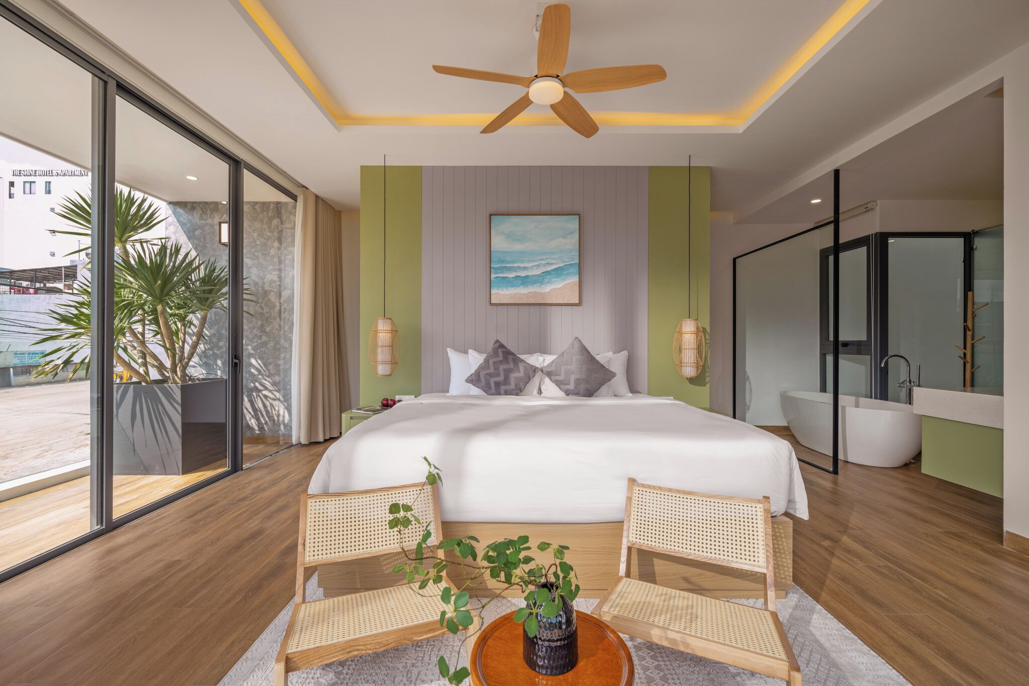 A modern hotel room with a large bed, two wicker chairs, a round wooden table with a plant, a ceiling fan, and a large window offering a view of the outside.