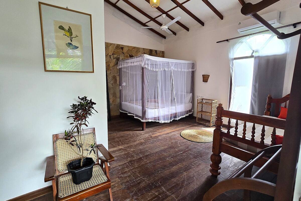 Una habitación con una cama bajo un dosel blanco, una silla de madera, un sillón de mimbre y una mesita con una planta.