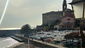 A marina with numerous boats docked along a stone quay, a building with a clock tower in the background, and a clear view of the sea.