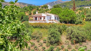 Une maison blanche avec un toit en tuiles, entourée de verdure et avec des collines en arrière-plan.