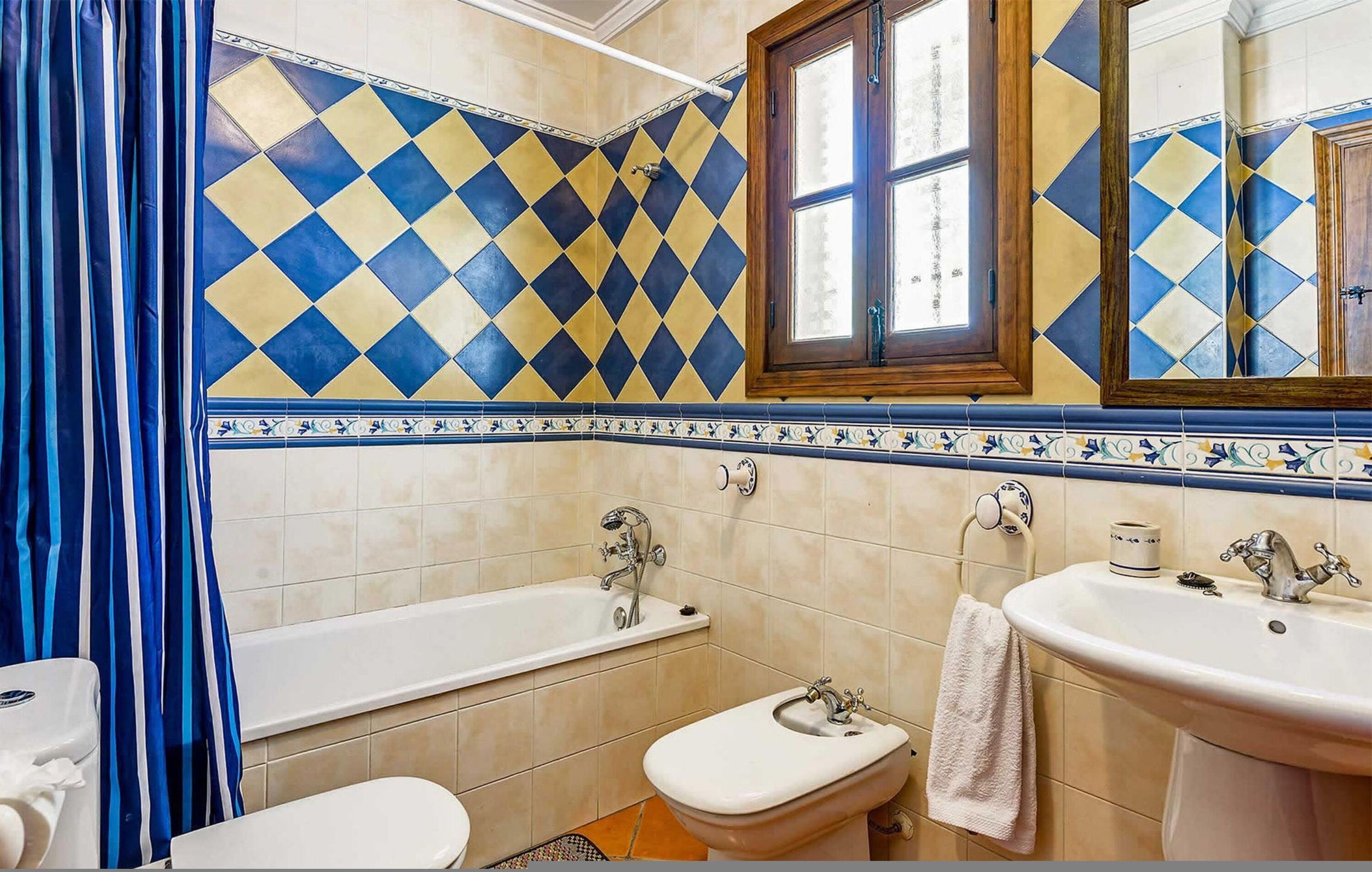 Baño con bañera, inodoro y lavamanos, con azulejos de diseño de diamantes en tonos azul y amarillo y una ventana con marco de madera.