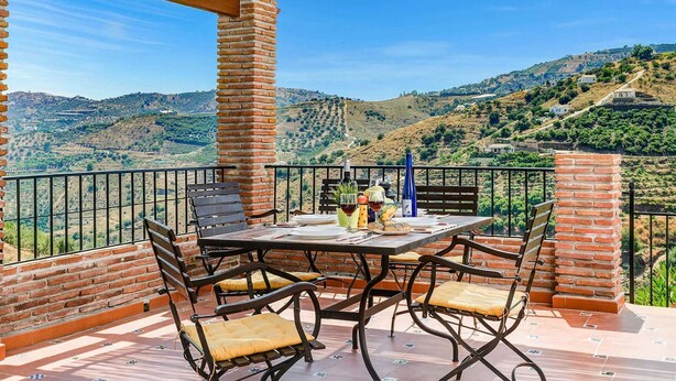 Une terrasse avec une table dressée pour le dîner, entourée de piliers en briques, d’où l’on peut admirer un paysage de collines verdoyantes et de vignobles.
