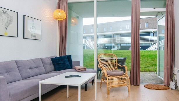Ein modernes Wohnzimmer mit grauem Sofa, einem weißen Couchtisch, einem Korbstuhl und einer Glastür, die auf einen Balkon mit Blick auf ein Gebäude und Grünflächen führt.