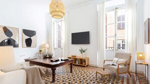 Ein modernes Wohnzimmer mit einem geometrisch gemusterten Boden, einem Holzesstisch, einer Couch, einem Sessel und einem an der Wand befestigten Fernseher.