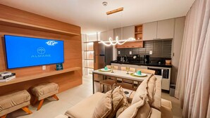 A modern apartment with a flat-screen TV displaying the Almaré logo, a dining area with a table and chairs, a kitchenette with a stove and microwave, and a sofa with cushions. - Flat FM20 Cabo Branco L6 - By Almare (João Pessoa)