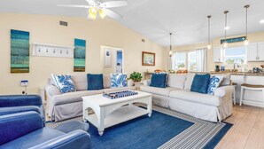 A living room with a sofa, coffee table, and blue chairs. There is a ceiling fan, a kitchen area with a window, and a dining table. - Life's A Beach - Pet Friendly, Private Pool, Beach Views, Across from Beach! (St George Island)